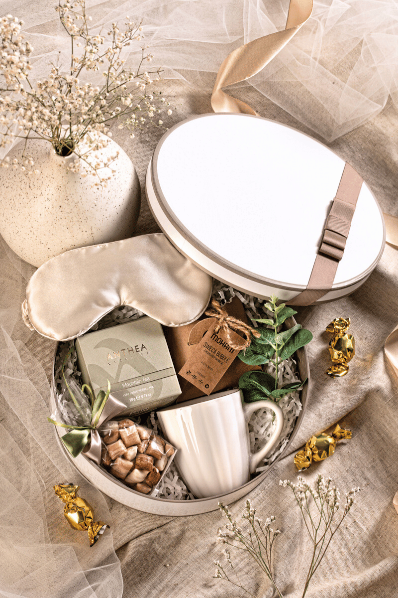 Otwarty, okrągły kremowy zestaw prezentowy z herbatą, kubkiem, świecą bąbelkową, satynową opaską i karmelkami. Idealny prezent na imieniny i relaks.