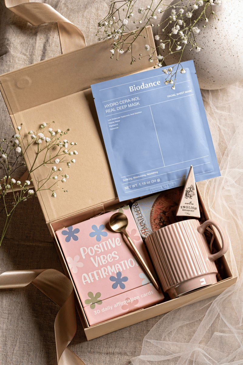 Otwarty beżowy zestaw prezentowy „Pozytywne Pudełko” z kartami, maską Biodance, czekoladkami i różowym kubkiem dla self-care.