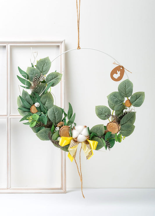 Ręcznie wykonany wianek wielkanocny z zielonymi listkami, piórkami, jajeczkami, drewienkami i kwiatem bawełny, zawieszony na drzwiach.