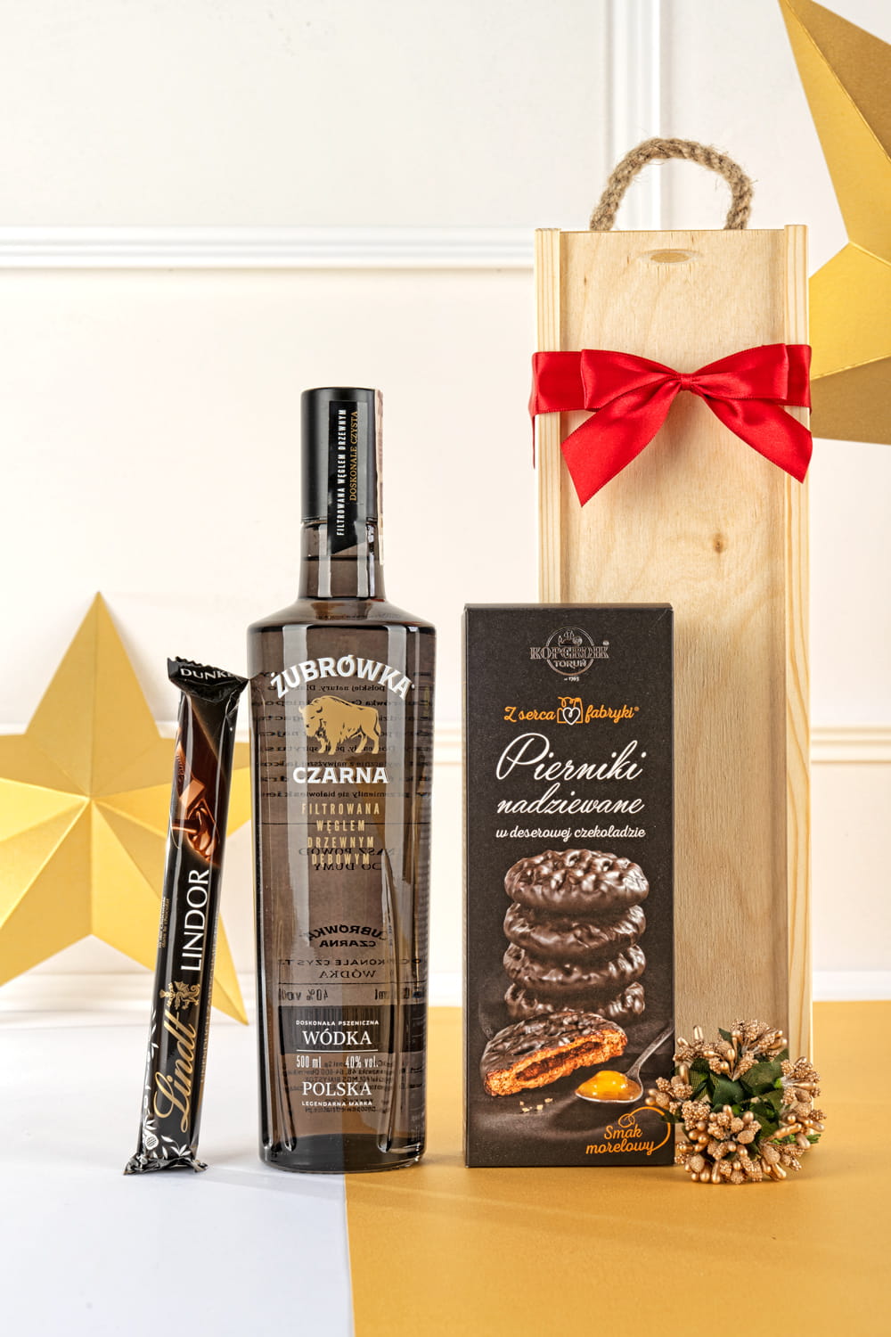 Luksusowy świąteczny zestaw prezentowy w otwartej drewnianej skrzyni z Żubrówką Czarną, czekoladą Lindt i piernikami, przewiązany czerwoną wstążką.