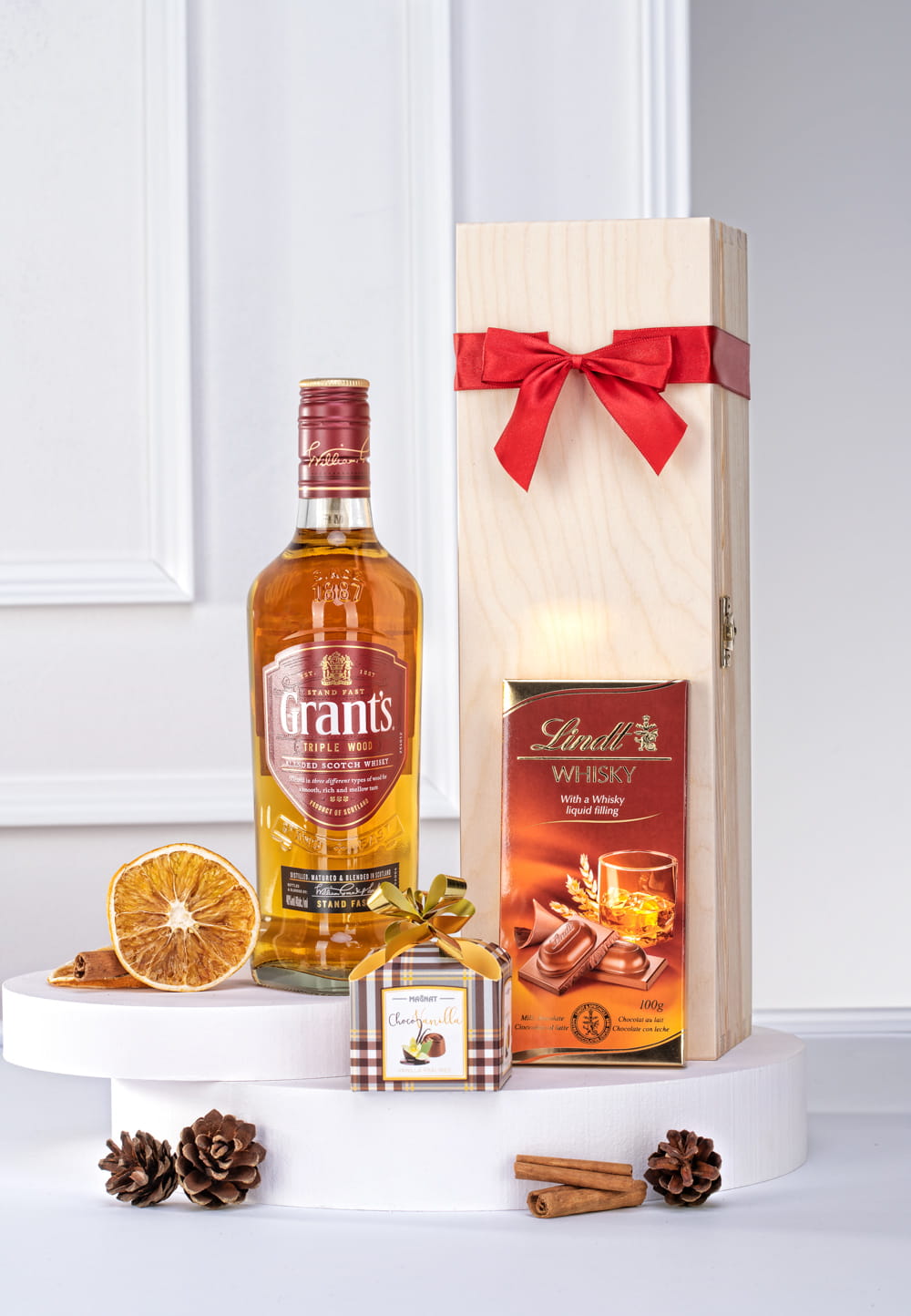 Elegancki zestaw świąteczny: whisky Grant's, czekolada Lindt, pistacjowe praliny w drewnianej skrzynce z czerwoną kokardą.