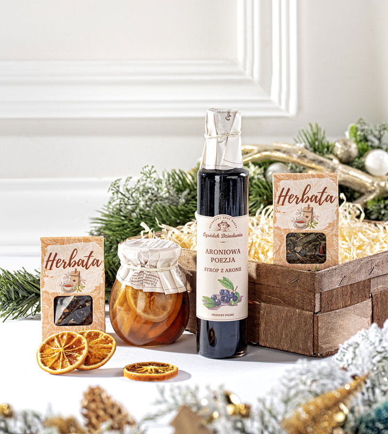 Elegancki kosz prezentowy z syropem z aronii, cytrynkami z cynamonem i dwoma ozdobnymi pudełkami herbat liściastych.