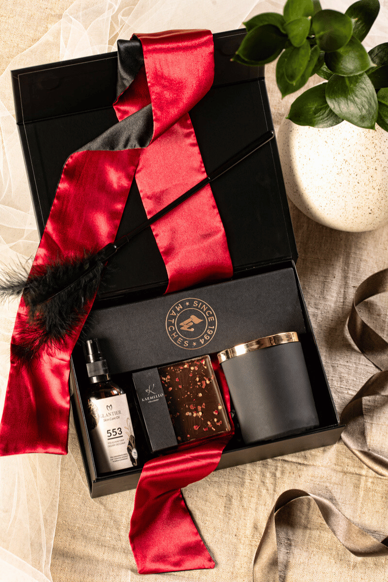 Zmysłowy zestaw prezentowy dla dwojga: otwarte czarne pudełko z satynową opaską, świecą, olejkiem do masażu i czekoladą.