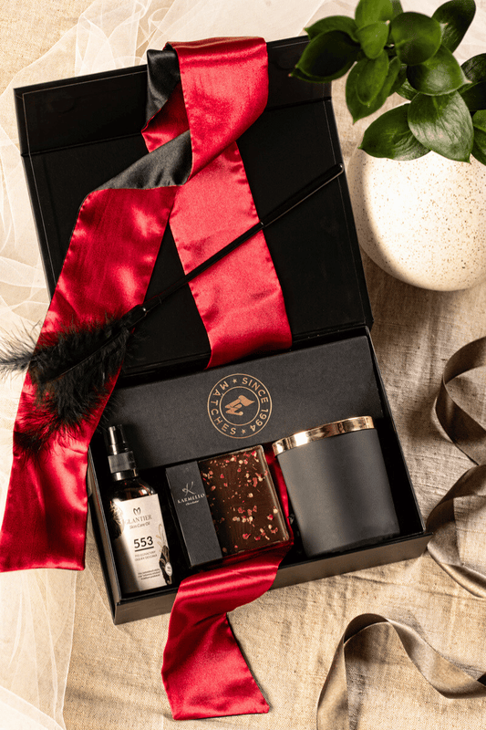 Zmysłowy zestaw prezentowy dla dwojga: otwarte czarne pudełko z satynową opaską, świecą, olejkiem do masażu i czekoladą.